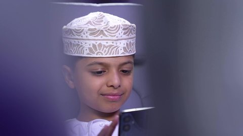 Using advanced technology and modern techniques, the concept of tourism and travel, preparing for an enjoyable summer vacation, a close-up of an Arab Gulf Omani child traveler wearing a dishdasha and a sleeve, smiling, sitting in an airplane seat and browsing a mobile phone, a tourist trip through Oman Air