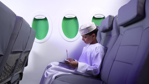ايماءات السعادة و السرور، مفهوم السياحة و السفر،الاستعداد لقضاء عطلة صيفية ممتعة، تصوير لطفل مسافر عربي خليجي عماني يرتدي الدشداشة و الكمه مبتسم يجلس في مقعد الطائرة و يقوم برسم شيء ما، رحلة سياحية من خلال خطوط الطيران العماني