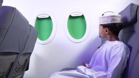 الاستعداد لقضاء عطلة صيفية ممتعة، تصوير لطفل مسافر عربي خليجي عماني يرتدي الدشداشة و الكمه مبتسم يجلس في مقعد الطائرة، رحلة سياحية من خلال خطوط الطيران العماني، ايماءات السعادة و السرور