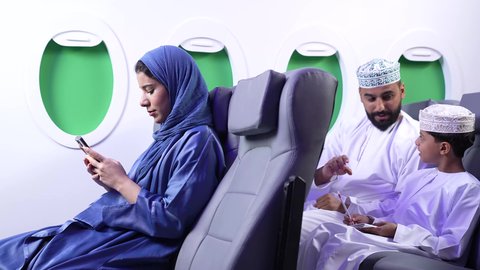 مفهوم السياحة والسفر، قضاء أوقات ممتعة في الإجازة، تصوير لعائله عربيه خليجيه عمانية يرتدون الزي التقليدي داخل الطائرة ، امراة خليجيه تقوم بامساك الهاتف المحمول ، الطيران العماني الخليجي