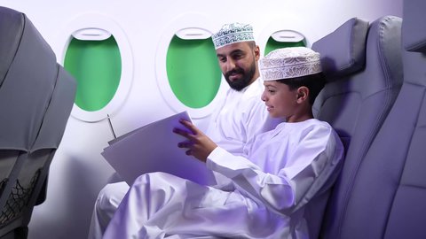 ايماءات السعادة و السرور، الطيران العماني الخليجي، مفهوم ممارسه الانشطه والعمل عن بعد داخل الطائره, تصويرلرجل عربي خليجي عماني يرتدي الدشداشه والكمه يجلس على كرسيه داخل الطائره الخليجيه يقوم بالعمل على جهاز الحاسوب المحمول برفقة ولده