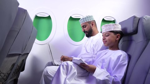 الطيران العماني الخليجي، مفهوم ممارسه الانشطه والعمل عن بعد داخل الطائره, تصويرلرجل عربي خليجي عماني يرتدي الدشداشه والكمه يجلس على كرسيه داخل الطائره الخليجيه يقوم بالعمل على جهاز الحاسوب المحمول برفقة ولده