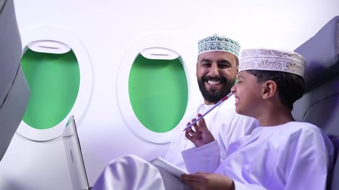 استخدام التكنولوجيا و التقنية ، خطوط الطيران العماني، رحلة سياحية عائلية، طفل يقضي وقت الرحلة مع والده، تصوير لرجل عربي خليجي عماني يرتدي الدشداشة و الكمه برفقة ولده  يمسك الحاسب المحمول في الطائرة، قضاء العطلة الصيفية برفقة العائلة