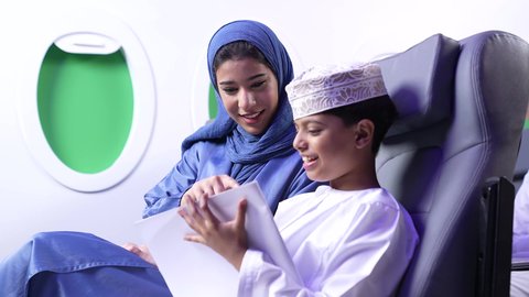 رحلة سياحية عائلية، طفل يقضي وقت الرحلة في مشاهده الرسومات،تصوير لامرأة عربية عمانية خليجية برفقة ولدها في الطائرة، قضاء العطلة الصيفية برفقة العائلة، خطوط الطيران العماني
