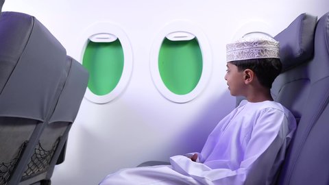 الاستعداد لقضاء عطلة صيفية ممتعة، تصوير من الجانب لطفل مسافر عربي خليجي عماني يرتدي الدشداشة و الكمه مبتسم يجلس في مقعد الطائرة، رحلة سياحية من خلال خطوط الطيران العماني، ايماءات السعادة و السرور
