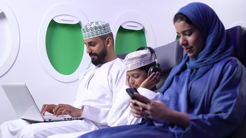 استخدام السياحة في إجازة الصيف, دمج التقنيات الحديثه فى الحياه اليوميه ، تصوير لعائله عربيه خليجيه عمانية يرتدون الزي التقليدي يجلسون في الطائرة ، امراة خليجيه ترتدي العباءة الزرقاء تقوم بامساك الجوال، ايماءات السعادة و السرور