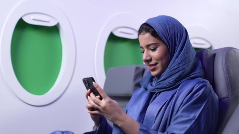 استخدام السياحة في إجازة الصيف, دمج التقنيات الحديثه فى الحياه اليوميه ، تصوير مقرب لامراة عربيه خليجيه عمانية ترتدي العباءة الزرقاء تجلس على كرسي داخل الطائره تمسك بيدها الهاتف المحمول