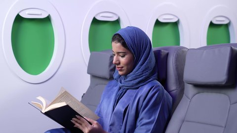 الترفيه والتسليه وقضاء الاوقات داخل الطائره، الاستمتاع بالقراءه والثقافه ، ممارسه الهوايات المفضله ، تصوير مقرب لامرأة عربية خليجية عمانية ترتدي العباءة الزرقاء تقوم بقراءة كتاب ما على كرسي الطائره في رحله جويه