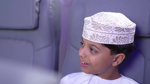 الاستعداد لقضاء عطلة صيفية ممتعة، تصوير مقرب لطفل مسافر عربي خليجي عماني يرتدي الدشداشة و الكمه مبتسم يجلس في مقعد الطائرة، رحلة سياحية من خلال خطوط الطيران العماني، ايماءات السعادة و السرور