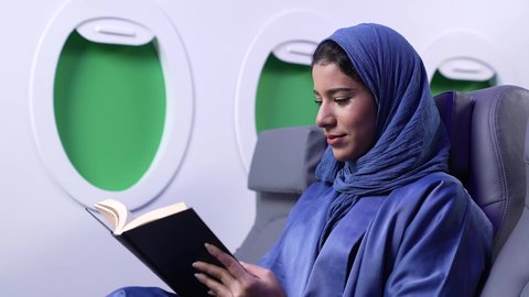 الاستمتاع بالقراءه والثقافه ، ممارسه الهوايات المفضله, الترفيه والتسليه وقضاء الاوقات داخل الطائره ، تصوير مقرب لامرأة عربية خليجية عمانية ترتدي العباءة الزرقاء تقوم بقراءة كتاب ما على كرسي الطائره في رحله جويه