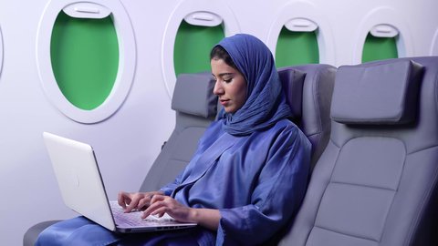 الطيران العماني الخليجي، مفهوم ممارسه الانشطه والعمل عن بعد داخل الطائره, تصوير لامرأة عربية خليجية عمانية ترتدي العباءة الزرقاء تقوم بالعمل على الحاسب المحمول داخل الطائرة