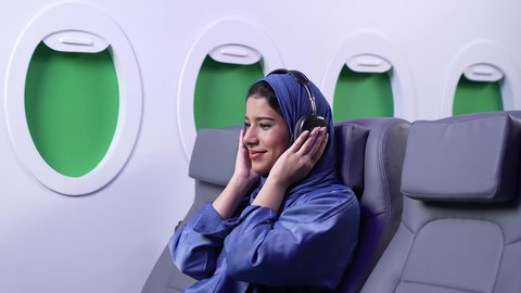 الاستماع للأغاني باستخدام الهيدفون، استخدام البرامج الصوتية في الجوال، تتصوير مقرب لامراة عربيه خليجيه اماراتية ترتدي العباءة الزرقاء  مبتسمة تضع سماعة الرأس  داخل الطائرة ، الطيران العماني الخليجي