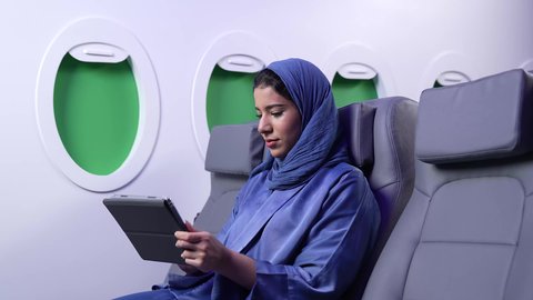 دمج التقنية والتكنولوجيا الحديثة في الحياة اليومية، تصويرلامرأة عربية خليجية عمانية ترتدي العباءة الزرقاء تقوم بالنقر على التابلت داخل الطائرة ، تصفح مواقع ووسائل التواصل الاجتماعي، شابة سعودية تعمل على الجهاز اللوحي، الاستمتاع باجازه الصيف