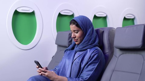استخدام السياحة في إجازة الصيف, دمج التقنيات الحديثه فى الحياه اليوميه ، تصوير مقرب لامراة عربيه خليجيه عمانيه ترتدي العباءه السوداء تجلس على كرسي داخل الطائره تمسك بيدها الهاتق المحمول و تنظر الى الكاميرا بايماءات السعادة و السرور