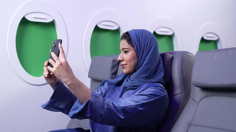 Using modern and advanced technological techniques, taking a souvenir selfie, gestures of happiness and pleasure, a close-up of an Omani Gulf Arab woman wearing a blue abaya taking a selfie on a mobile phone inside the plane, a tourist trip through Oman Air
