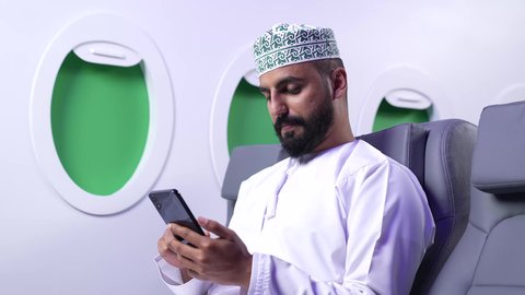 استخدام السياحة في إجازة الصيف, دمج التقنيات الحديثه فى الحياه اليوميه ، تصوير مقرب لشاب عربي خليجي عماني يرتدي الدشداشه والكمه يجلس على كرسيه داخل الطائره ويمسك بيده الهاتف المحمول
