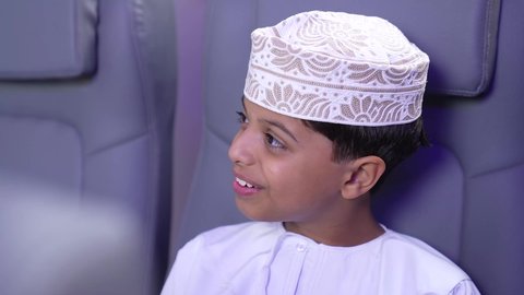 Preparing for a fun summer vacation, a close-up of an Arab Gulf Omani child traveler wearing a dishdasha and a keffiyeh, smiling, sitting in an airplane seat, a tourist trip with Oman Air, gestures of happiness and pleasure