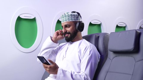 الاستماع للأغاني باستخدام الهيدفون، استخدام البرامج الصوتية في الجوال، تصوير  لرجل عربي خليجي عماني مبتسم يرتدي الدشداشة و الكمة  يضع سماعة الرأس يمسك بيده الهاتف المحمول داخل الطائرة ، الطيران العماني الخليجي