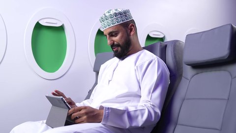 استخدام التكنولوجيا والتقنيات الحديثة, تصفح مواقع التواصل الاجتماعي، متابعة الأفلام والمسلسلات الممتعة، المحادثة مع الأهل والأصدقاء، تصوير مقرب لرجل عربي خليجي عماني يرتدي الدشداشة و الكمة يحمل بيده جهاز التابلت داخل الطائرة، الطيران العماني الخليجي