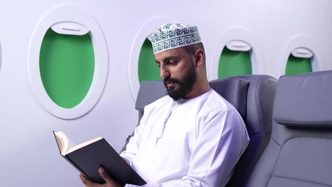 الطيران العماني الخليجي، ممارسه الانشطه الترفيهيه وهوايه المطالعه, تصوير مقرب من الجانب لرجل عربي خليجي عماني يرتدي الدشداشه والكمه يجلس على كرسيه داخل الطائره يقرأ كتابا بيده