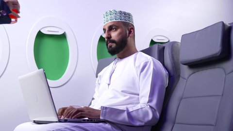 مفهوم ممارسه الانشطه والعمل عن بعد داخل الطائره, تصوير مقرب من الجانب لرجل عربي خليجي عماني يرتدي الدشداشه والكمه يجلس على كرسيه داخل الطائره الخليجيه يقوم بالعمل على جهاز الحاسوب المحمول, الطيران العماني الخليجي