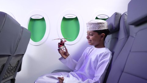 الاستعداد لقضاء عطلة صيفية ممتعة، مفهوم السياحة و السفر، ايماءات السعادة و السرور، تصوير من الجانب لطفل مسافر عربي خليجي عماني يرتدي الدشداشة و الكمه ويجلس في مقعد الطائرة يقوم بامساك طائرة لعبه، رحلة سياحية من خلال خطوط الطيران العماني