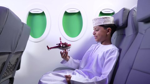 مفهوم السياحة و السفر، ايماءات السعادة و السرور، الاستعداد لقضاء عطلة صيفية ممتعة، تصوير من الجانب لطفل مسافر عربي خليجي عماني يرتدي الدشداشة و الكمه ويجلس في مقعد الطائرة يقوم بامساك طائرة لعبه، رحلة سياحية من خلال خطوط الطيران العماني