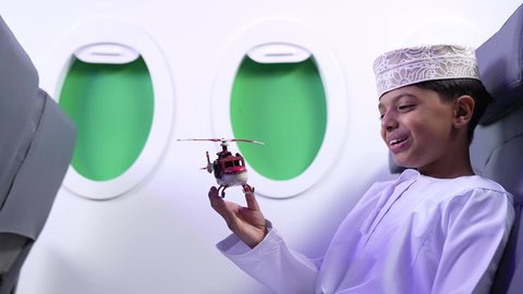 ايماءات السعادة و السرور، الاستعداد لقضاء عطلة صيفية ممتعة، تصوير مقرب من الجانب لطفل مسافر عربي خليجي عماني يرتدي الدشداشة و الكمه ويجلس في مقعد الطائرة يقوم بامساك طائرة لعبه، رحلة سياحية من خلال خطوط الطيران العماني