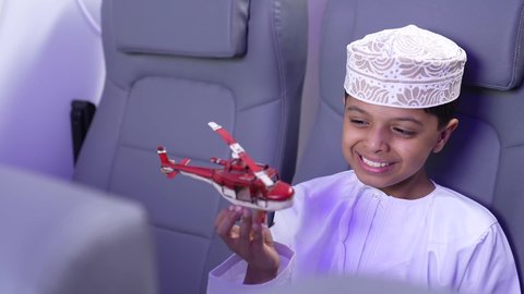 الاستعداد لقضاء عطلة صيفية ممتعة، تصوير مقرب لطفل مسافر عربي خليجي عماني يرتدي الدشداشة و الكمه ويجلس في مقعد الطائرة يقوم بامساك طائرة لعبه، رحلة سياحية من خلال خطوط الطيران العماني، ايماءات السعادة و السرور