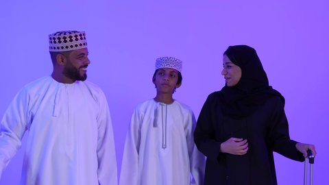 الذهاب في رحلة سفر ممتعة، الإستعداد لقضاء الإجازة الصيفية ، تصوير لرجل عربي خليجي عماني يرتدي الدشداشة والكمة يقف مع زوجته وابنه ويمسك حقيبة الأمتعة، الأجواء العائلية المليئة بالحب