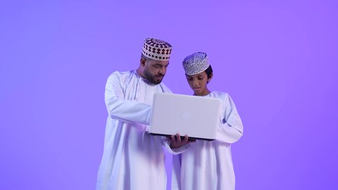 متابعة حصص الاونلاين عبر جهاز الحاسوب المحمول، أخبار جديده على مواقع التواصل الاجتماعي، تصوير لولد عربي خليجي عماني بجانب والده يقوم بالتعلم عن بعد بإيماءات الضحك و السعادة، تشجيع الأبناء والاعتناء بهم