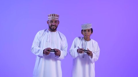 المتعة و التسلية بألعاب الفيديو، اللعب مع العائلة ، تصوير لأب وإبن عماني خليجي عربي يرتديان الدشداشة والكمة , يمسكان عصا التحكم و يلعبان بألعاب الفيديو، إيماءات وجه بالإندماج والتفاعل مع اللعبه