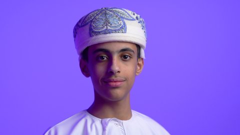 The concept of concern for elegance and outward appearance, expressions and gestures of pleasure and self-confidence, a close-up of an Arab Gulf Omani teenage boy wearing a dishdasha and turban looking at the camera with serious looks, blue background