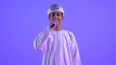 تقديم العروض الصوتية، تعابير الاندماج والتفاعل مع الموسيقى ، تصوير مقرب لولد مراهق عربي خليجي عماني يرتدي العمامة و الدشداشة يمسك بيده المايكروفون يقوم بالغناء ينظر الى الكاميرا بإيماءات الفرح والسرور، تسجيل مقطع صوتي غنائي، خلفية زرقاء