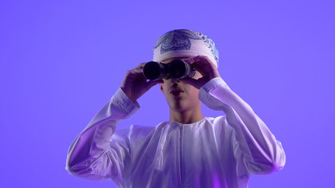 Seeing distant things, future planning for goals and ambitions, love of exploration and adventure, close-up of an Arab Gulf Omani teenage boy wearing a dishdasha and turban holding binoculars, thinking and looking to the future, blue background