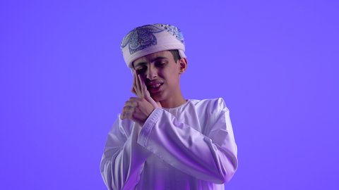 إيماءات الانزعاج والألم الشديد, مشاكل التهاب اللثة, طلب العلاج والمساعدة, الحاجة لزيارة طبيب الأسنان, بورتريه مقرب لولد عربي خليجي عماني يرتدي الدشداشة و الكمة يضع يده على خده, خلفية زرقاء .