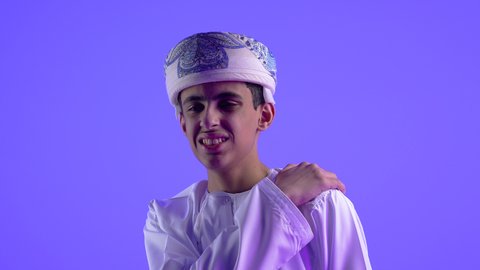 التهاب العضلات والمفاصل، الإحساس بالتعب والإرهاق, إيماءات الألم في الأكتاف، بورتريه مقرب لولد عربي خليجي عماني يرتدي الدشداشة والكمة يضع يده على كتفه بتعابير الوجع، الحاجه لوضع خطة علاجية, طلب المساعده للذهاب الى المستشفى، خلفية زرقاء .