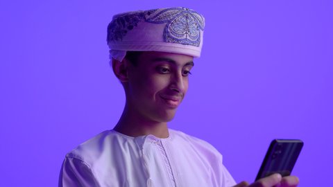 تبادل الأحاديث مع الأهل والأصدقاء، استخدام التكنولوجيا والأجهزة التقنية الحديثة المتطورة، ايماءات السعادة والفرح، تصوير مقرب لولد مراهق عربي خليجي عماني مبتسم يرتدي الدشداشة والعمامة يعمل على الهاتف المحمول، خلفية زرقاء