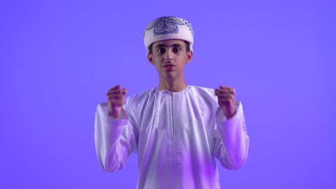 الانتصار والفوز والنجاح، مفهوم الانجاز وتحقيق الاهداف، تصوير مقرب لولد مراهق عربي خليجي عماني يرتدي الدشداشة والعمامة ينظر الى الكاميرا يرفع يديه للاعلى بإيماءات الفرح والسرور, الشعور بالسعادة والقوة, خلفية زرقاء