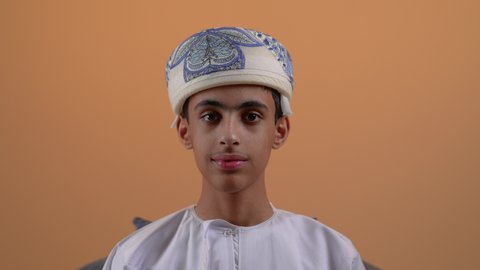 الاهتمام بالأناقة والمظهر الخارجي، تعابير وايماءات السرور والثقة بالنفس، تصوير مقرب لولد مراهق عربي خليجي عماني يرتدي الدشداشة والعمامة يجلس على كرسي مريح ينظر الى الكاميرا بنظرات الجدية، خلفية بيج