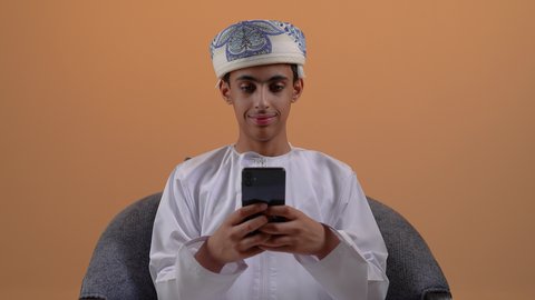 استخدام التكنولوجيا والأجهزة التقنية الحديثة المتطورة، تبادل الأحاديث مع الأهل والأصدقاء، ايماءات السعادة والفرح، تصوير مقرب لولد مراهق عربي خليجي عماني مبتسم يرتدي الدشداشة والعمامة يجلس على الكرسي يعمل على الهاتف المحمول، خلفية بيج