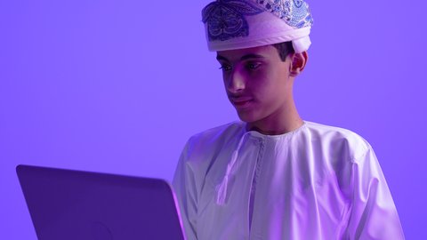 استخدام جهاز تقني حديث، دمج التكنولوجيا في الحياة، تصوير مقرب لولد مراهق عربي خليجي عماني يرتدي الدشداشة والعمامة يقوم بالعمل على الحاسب المحمول بتعابير وايماءات الاندماج والتركيز، خلفية زرقاء