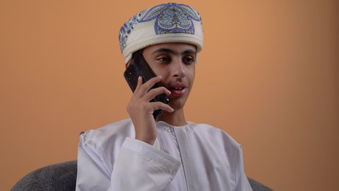 ايماءات السعادة و السرور، التواصل مع الاهل و الاصدقاء, تصوير مقرب لولد مراهق عربي خليجي عماني يرتدي الدشداشه والعمامة يجلس على كرسي مريح يقوم بإجراء مكالمة هاتفية عبر الهاتف المحمول، استخدام التقنيات الحديثة المتطورة, خلفية بيج
