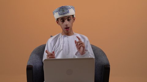 إجراء مكالمة فيديو وبث مباشر، استخدام جهاز تقني حديث، دمج التكنولوجيا في الحياة، تصوير مقرب لولد مراهق عربي خليجي عماني يرتدي الدشداشة والعمامة يقوم بالعمل على الحاسب المحمول بتعابير وايماءات الاندماج والتركيز، خلفية بيج