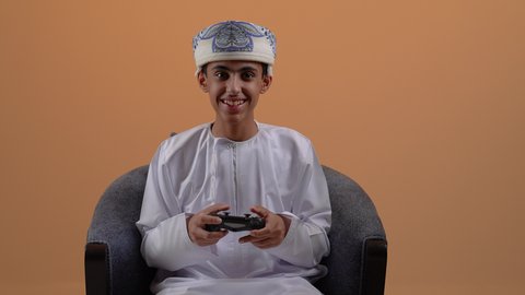 قضاء أوقات ممتعة في ألعاب الفيديو الترفيهيه، تصوير مقرب لولد مراهق عربي خليجي عماني يرتدي الدشداشة والعمامة يجلس على كرسي مريح يحمل بيده عصا التحكم، إيماءات الفرح والاستمتاع والاندماج، استخدام الأجهزة التكنولوجية والتقنيات الحديثة، خلفية بيج