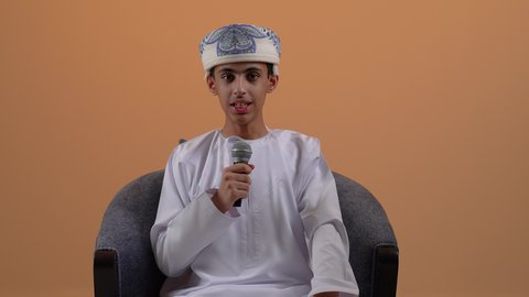 القاء الخطابات والحوارات، تقديم العروض الصوتية، تعابير الاندماج والتفاعل، تصوير مقرب لولد مراهق عربي خليجي عماني يرتدي العمامة و الدشداشة يمسك بيده المايكروفون يقوم بالتحدث ينظر الى الكاميرا بإيماءات الثقة بالنفس، تسجيل مقطع صوتي غنائي، خلفية بيج