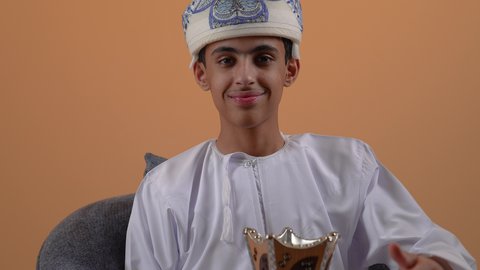 استخدام خشب العود بالأعياد والمناسبات, العادات والتقاليد العمانية، مفهوم التطيب بالبخور, تصوير مقرب لولد مراهق عربي خليجي عماني يرتدي الدشداشة والعمامة يجلس على كرسي مريح يقوم بالتعطر من المبخرة, الاهتمام بالمظهر الخارجي, خلفية بيج