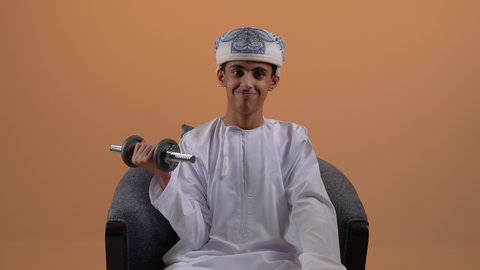 التدرب على رفع الأثقال والقيام بالتمارين الرياضية، تصوير مقرب لولد مراهق عربي خليجي عماني يرتدي الدشداشة والعمامة يقوم برفع الدامبلز، اتباع أسلوب حياة صحي، ممارسة الرياضة وبناء الكتلة العضلية،  خلفية بيج
