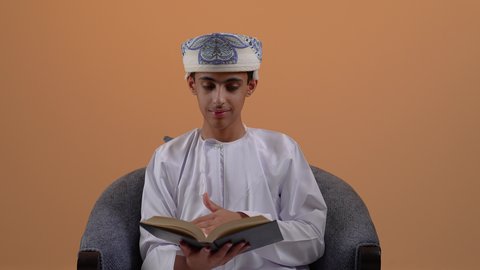 الاستمتاع بقراءة الكتب والقصص والروايات، قضاء الوقت بالتعلم والتثقف، ثقافة التعلم الذاتي، تصوير مرب لولد مراهق عربي خليجي عماني يرتدي الدشداشة والعمامة يجلس على الكرسي يحمل بيده كتاب، خلفية بيج