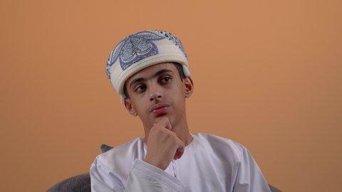 التمعن والتركيز و التخطيط للمستقبل، إيجاد الحل لمشكلة ما، تصوير مقرب لولد مراهق عربي خليجي عماني يرتدي الدشداشة والعمامة يضع يده تحت ذقنه بإيماءات التامل والتفكير، مفهوم شرود الذهن، خلفية بيج